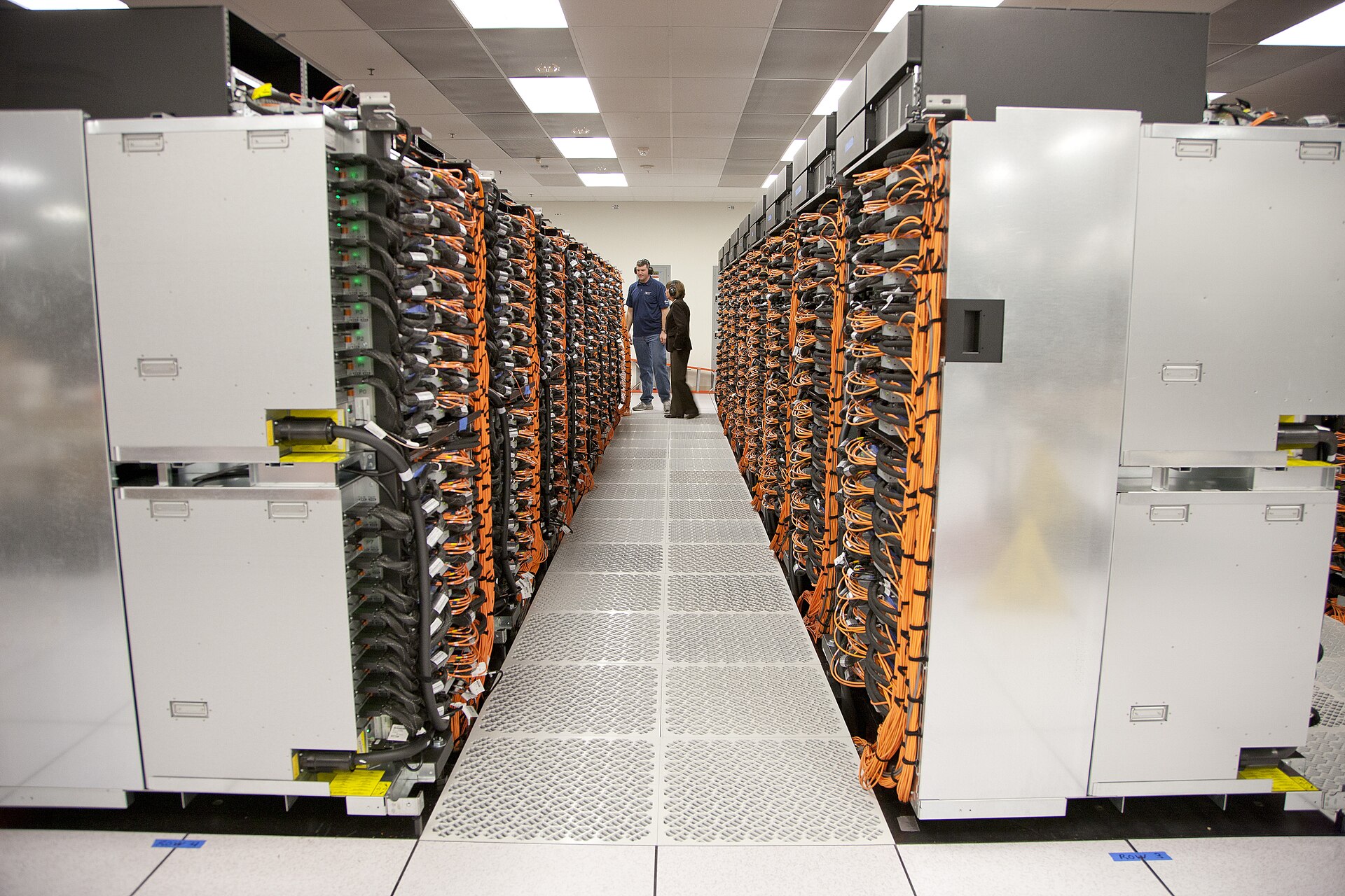A narrow datacenter aisle between two dense rows of supercomputer racks wired with orange interconnect cabling, with two technicians standing at the far end.