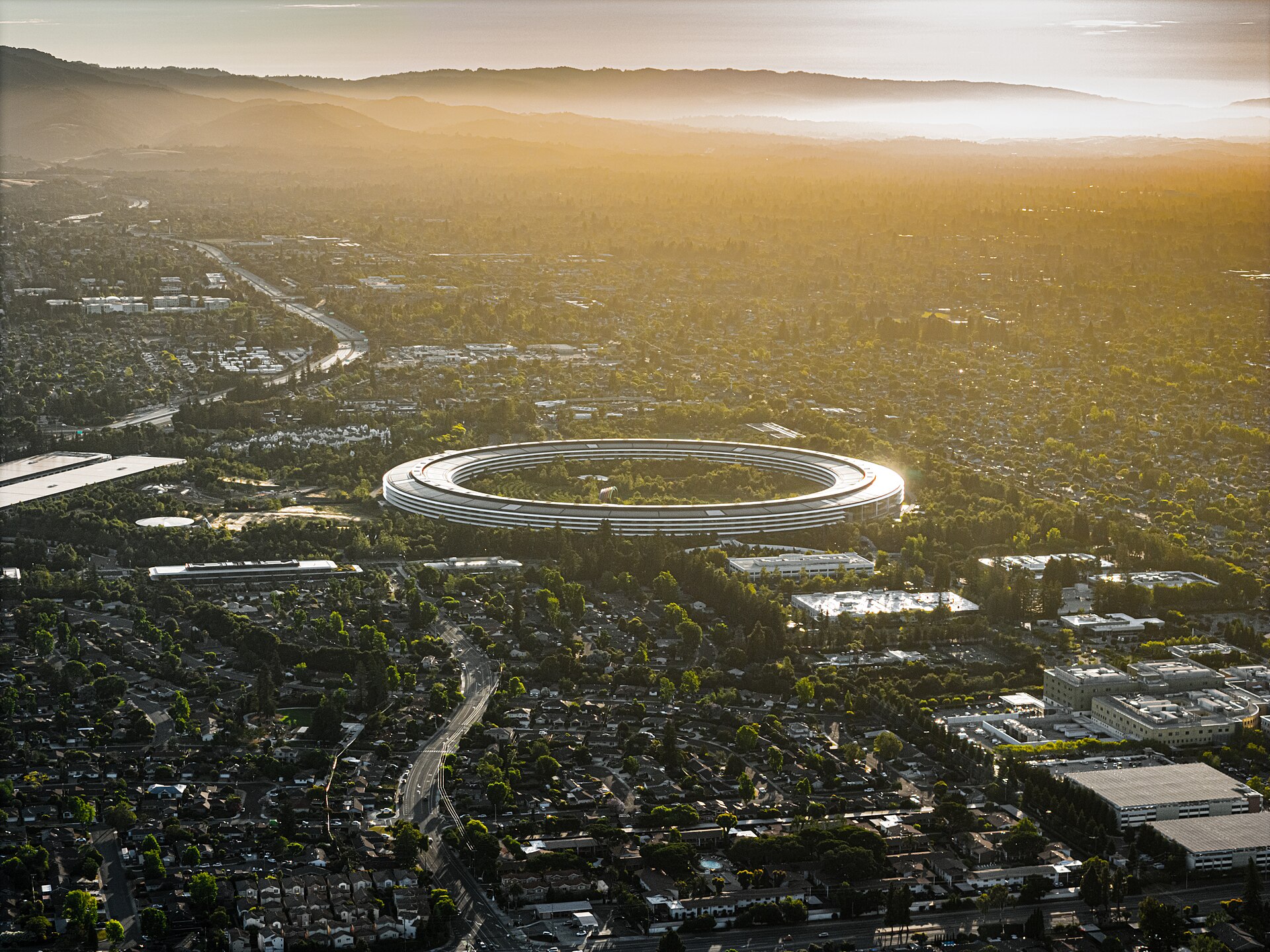 Aerial view of the Apple Park ring building in Cupertino at golden hour, with the surrounding Santa Clara Valley stretching toward hazy hills.
